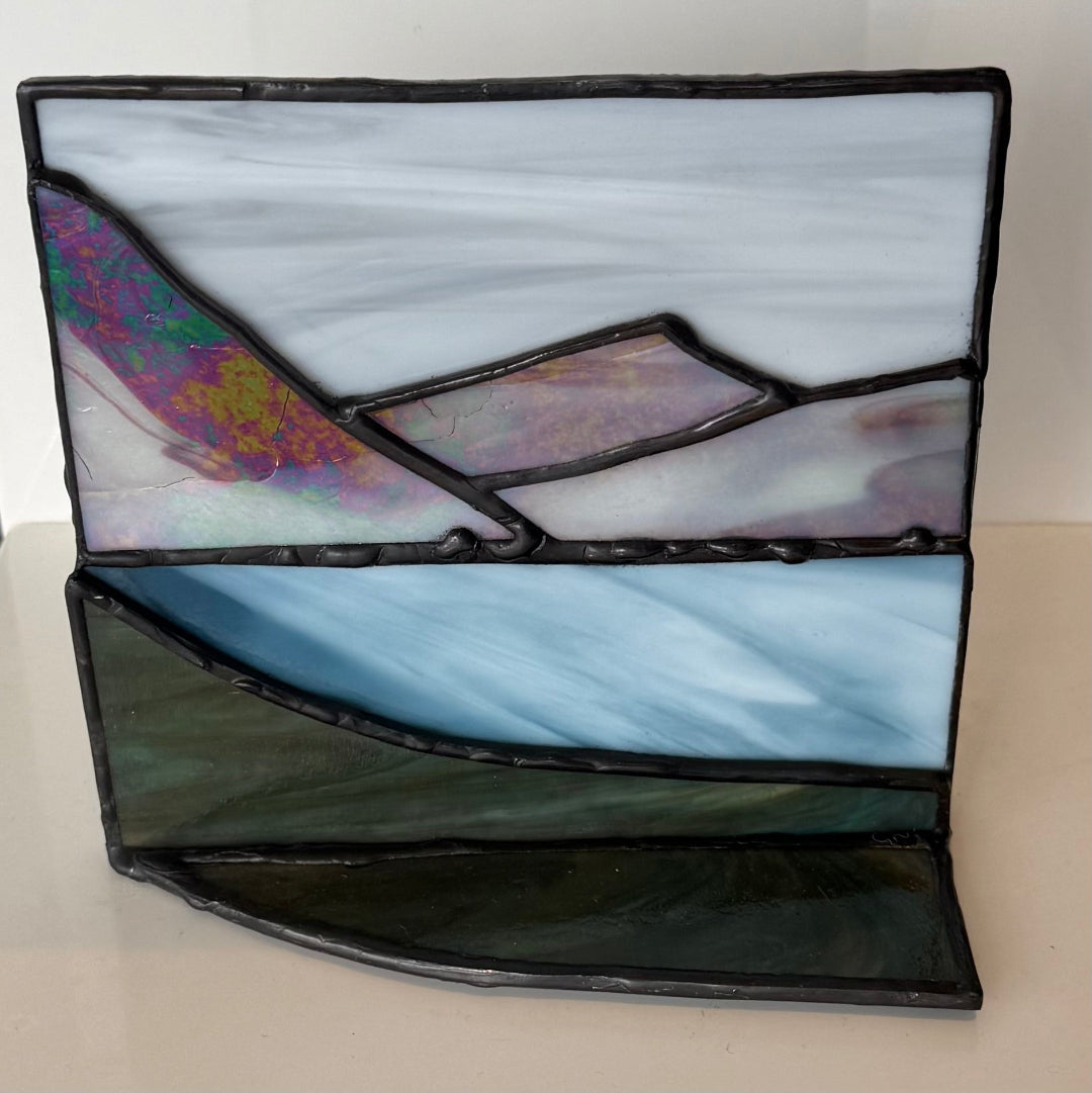 A stained glass panel depicting a landscape with a white sky, a blue sky, a dark green foreground, and an iridescent purple and gold section on the left, framed by black solder lines.