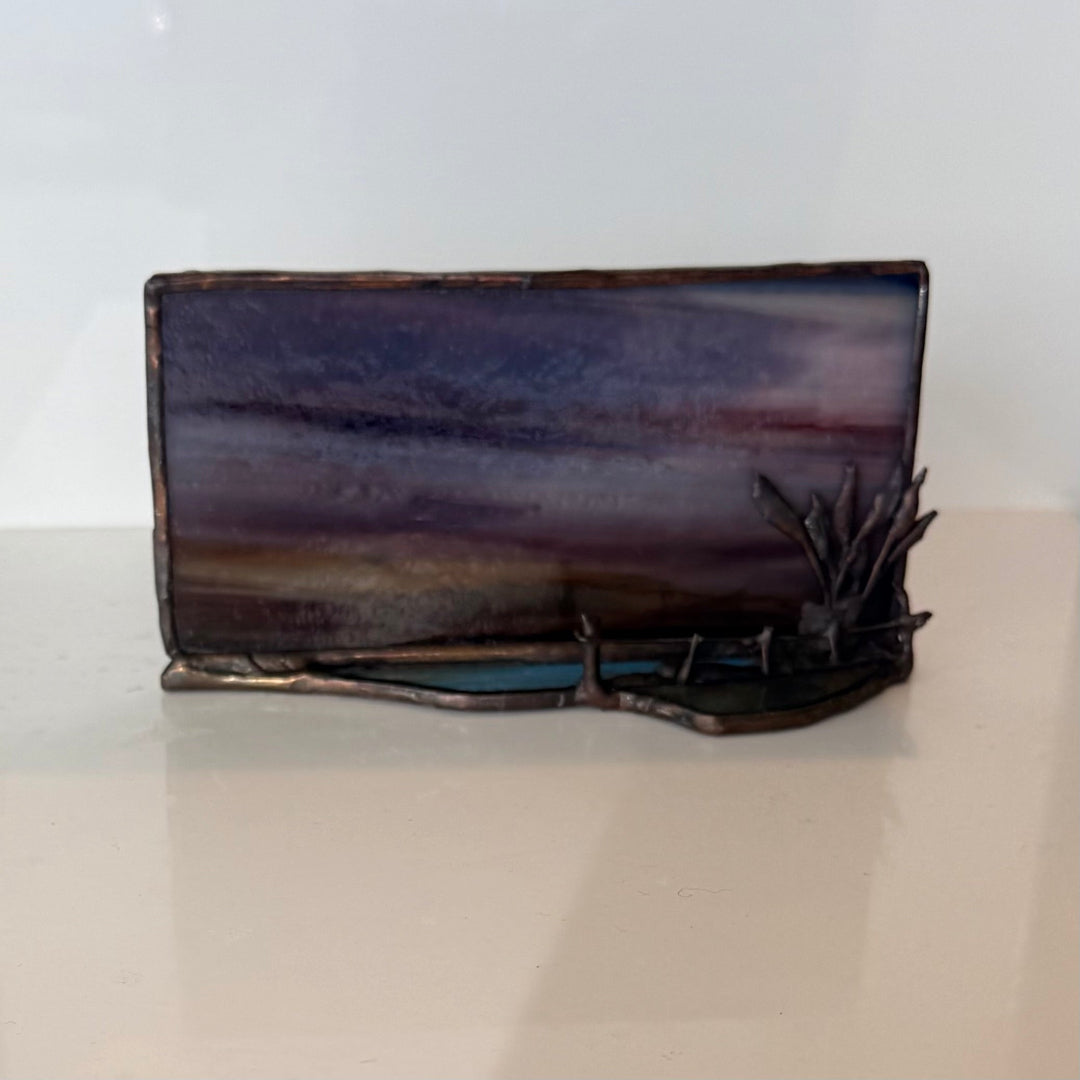 A rectangular stained glass art piece depicting a dark, moody landscape with purple, blue, and brown tones, featuring a small black metal reed sculpture on the right side, displayed on a light beige surface against a white background.