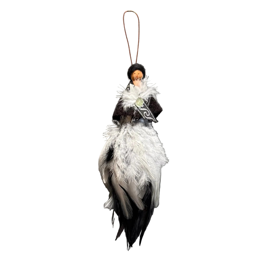 A handmade decorative ornament featuring a small peg doll with black hair and a face, dressed in a dark shawl with a white feather brooch, holding a silver-colored decorative object, and finished with a long white and black feather skirt hanging from a copper wire, set against a white background.