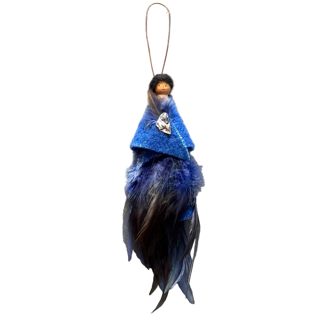 A handmade decorative hanging ornament featuring a small peg doll figure with black hair and a face, dressed in a royal blue felt shawl with a paua shell & feather brooch , and a blue-and-black feather skirt, with a copper wire loop suspended against a white background.