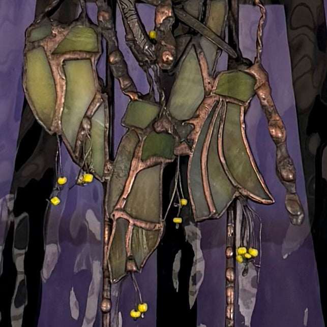A close-up of a stained-glass panel with green translucent kowhai flowers, copper-toned outlines, and small yellow bead accents against a dark purple background.