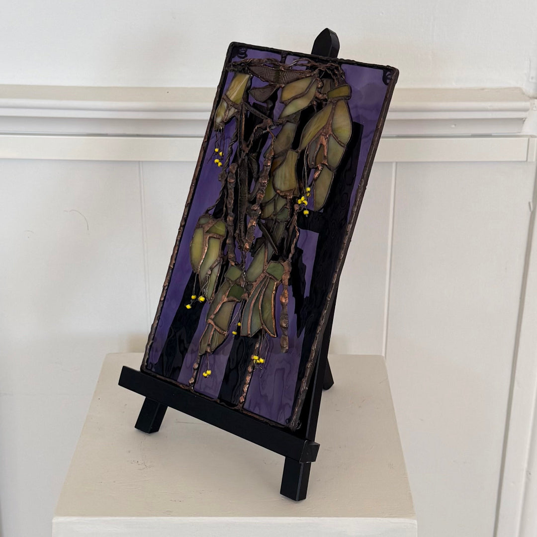 A stained glass panel depicting a cluster of kowhai flowers on a purple background, displayed on a black easel stand against a white wall.