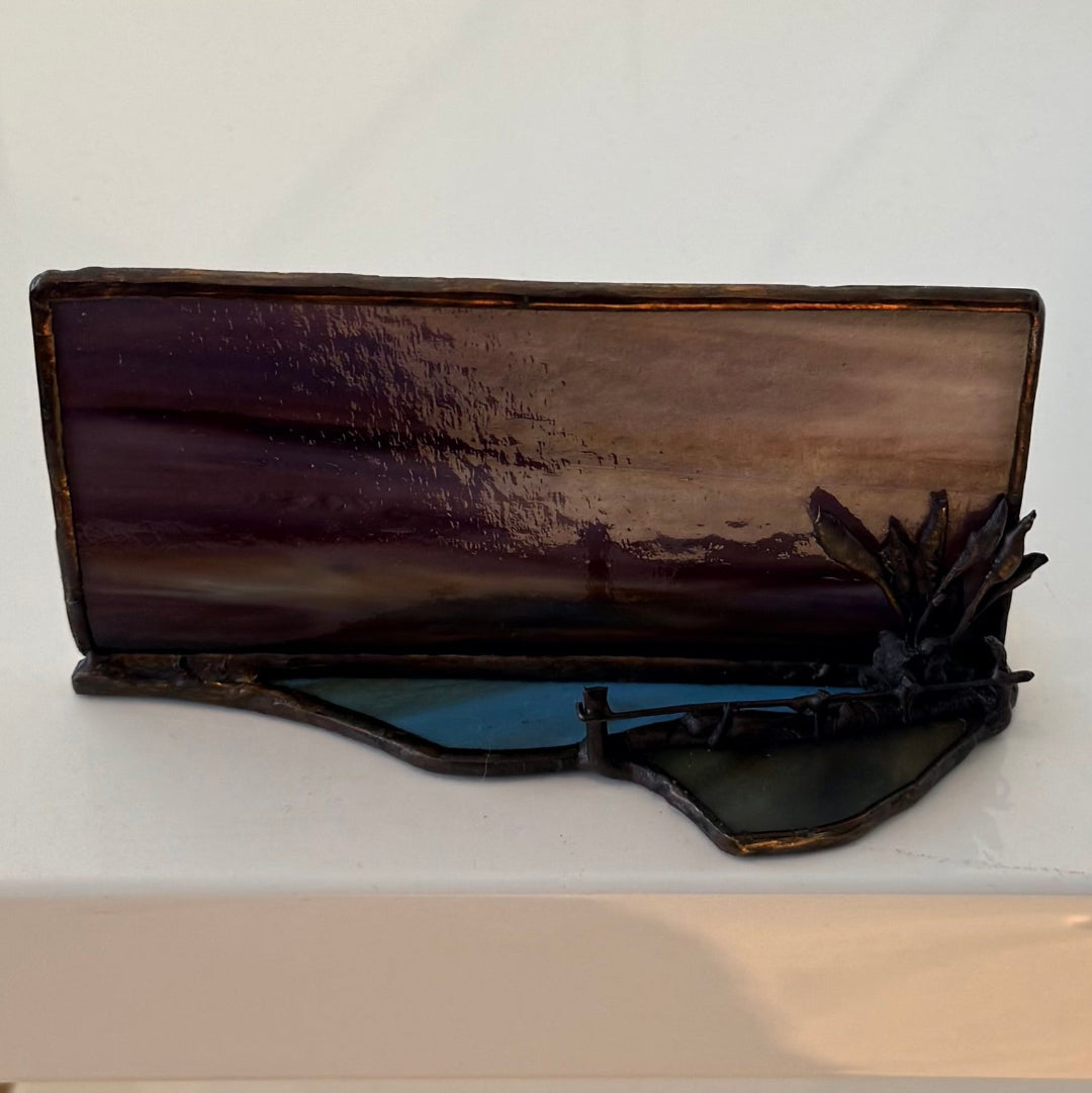 A rectangular stained glass art piece depicting a landscape scene with dark purple and brown tones, resting on a white surface.