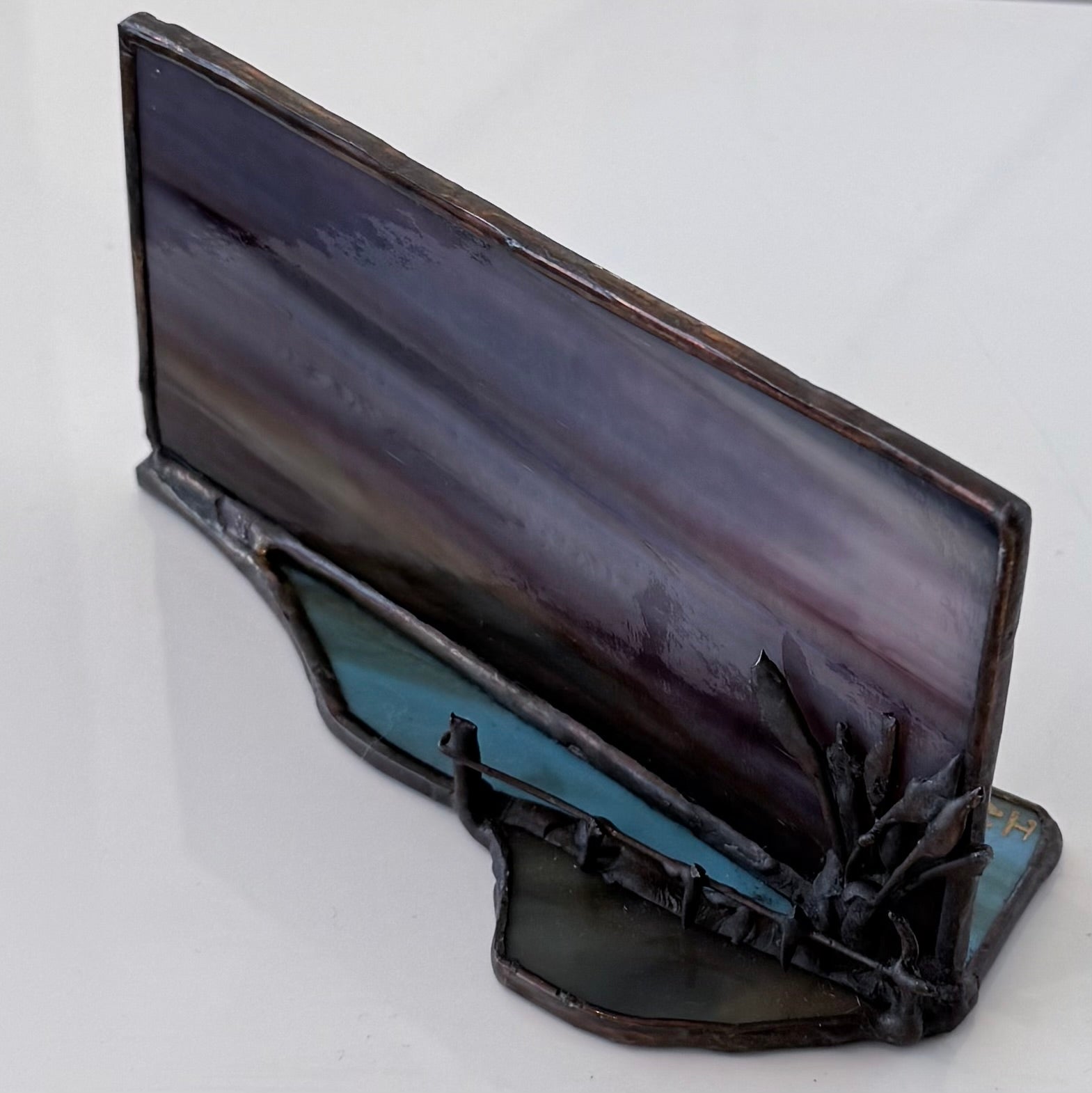 A stained glass art piece depicting a dark, moody landscape with a purple and gray sky, a teal-blue body of water, and a black rocky shoreline with small spires.