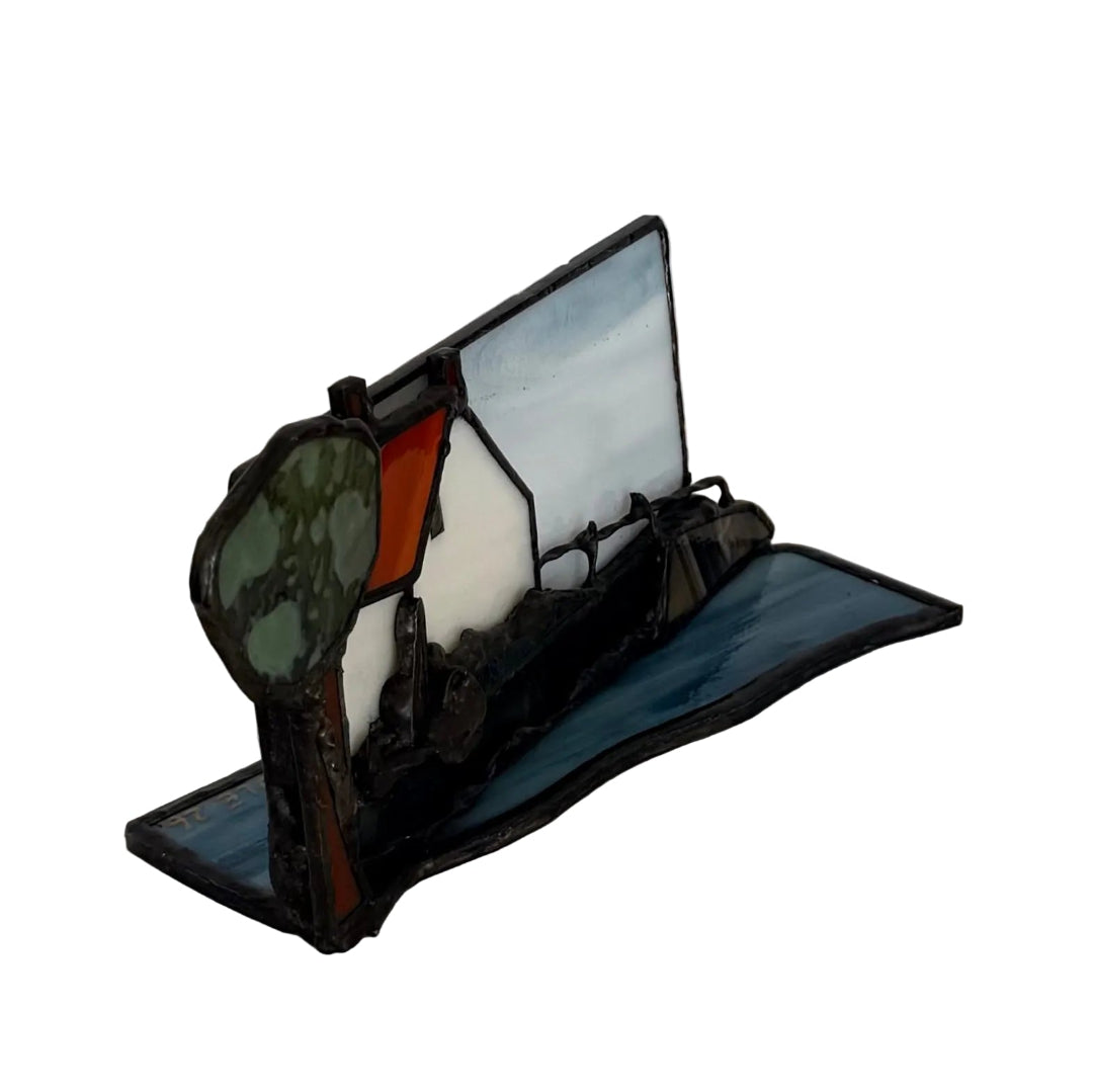 A stained glass sculpture featuring a coastal cttage in blue, white, green, and orange separated by dark metal framing, displayed on a light-colored surface against a plain wall.