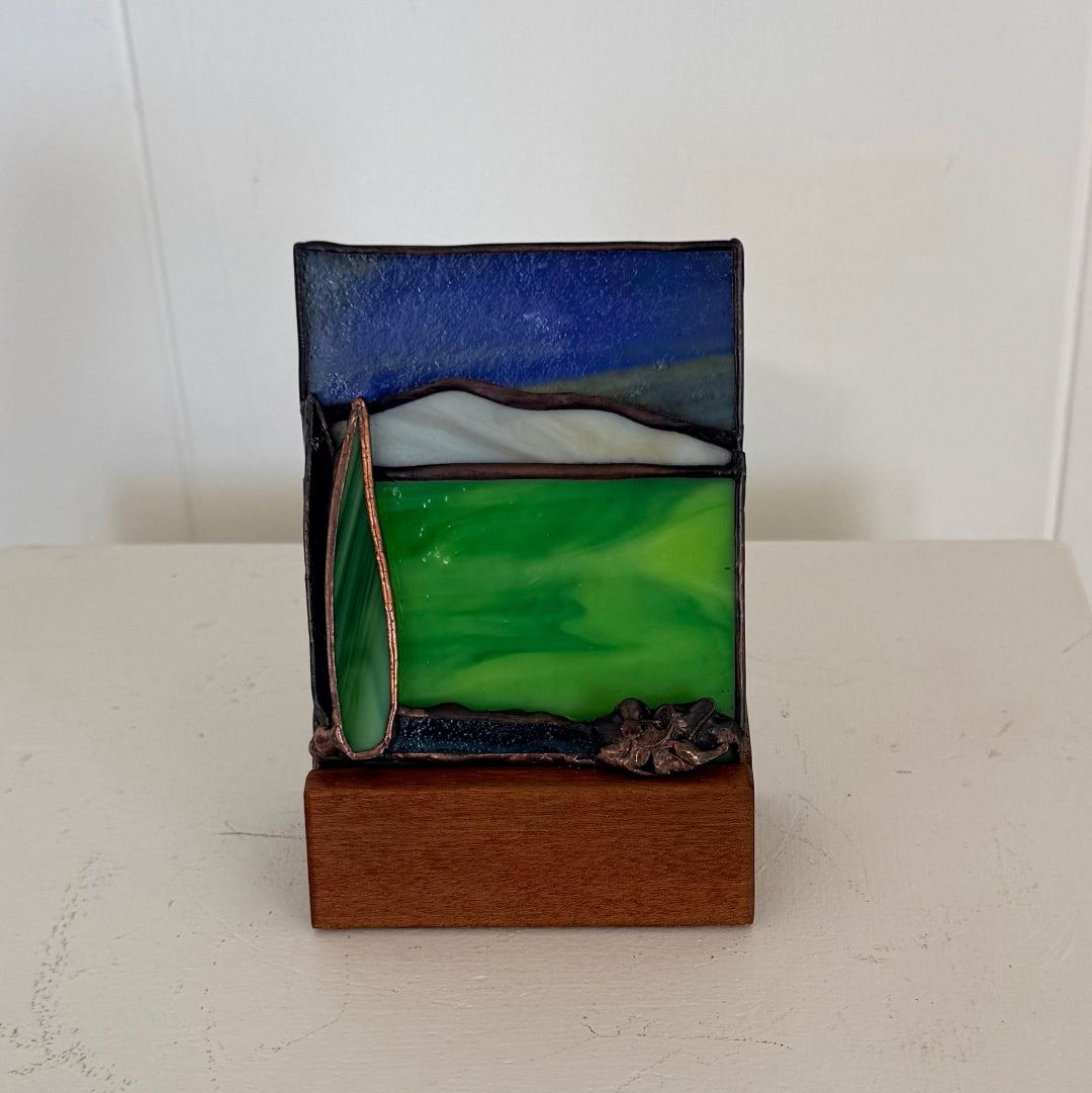 A small stained glass landscape sculpture on a wooden base, featuring a blue sky, green hills, and dark metallic outlines, set against a plain white background.