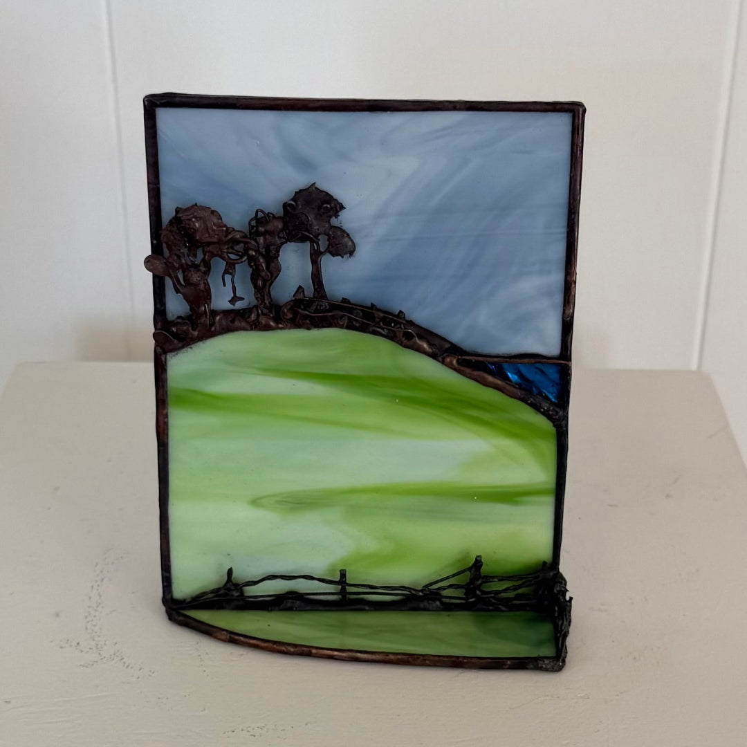 A rectangular stained glass art piece depicting a landscape with green fields, dark tree silhouettes, and a blue sky, set against a plain light background.