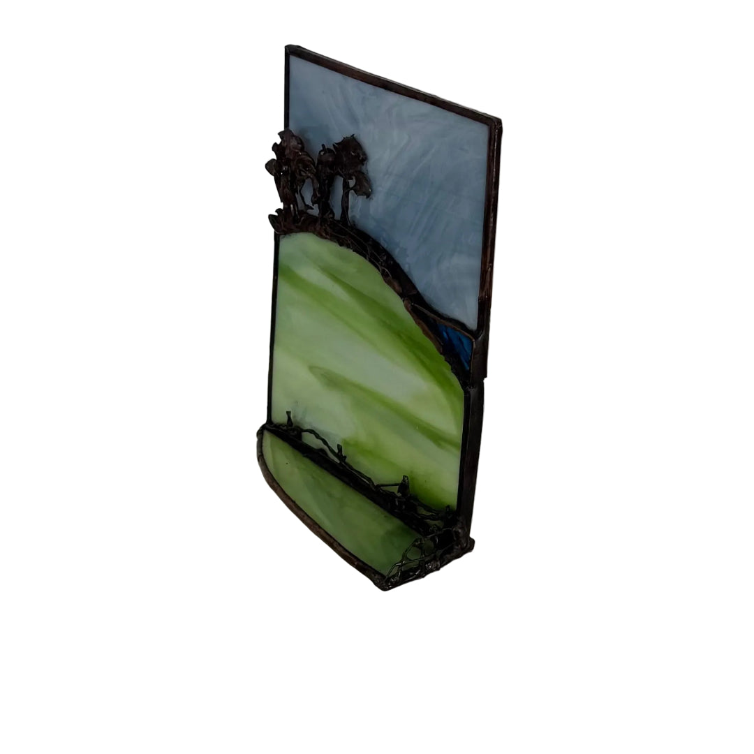 A stained glass landscape art piece featuring green hills, dark trees, and a blue sky, displayed on a white surface against a white paneled wall.