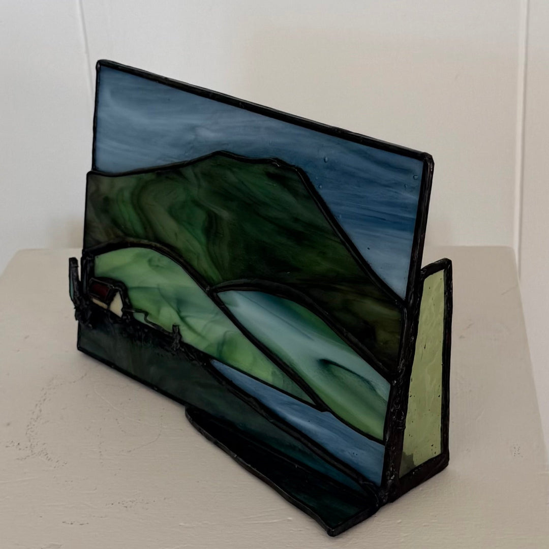 A stained glass art piece depicting a mountain landscape with green and blue tones, displayed on a light-colored surface against a white wall.