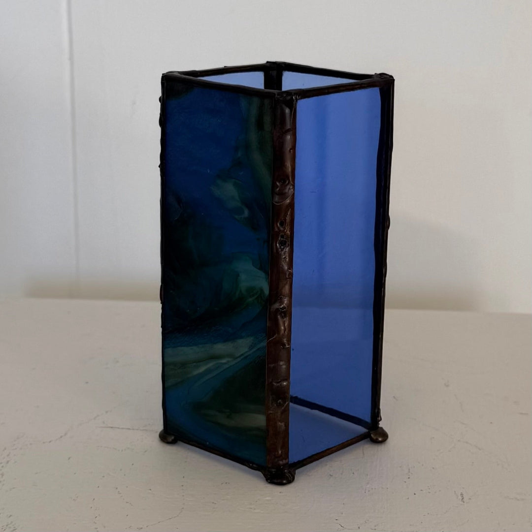 A small rectangular stained glass lantern with cobalt blue glass panels and a dark brown or black leaded metal frame, standing on a white surface against a white wall.
