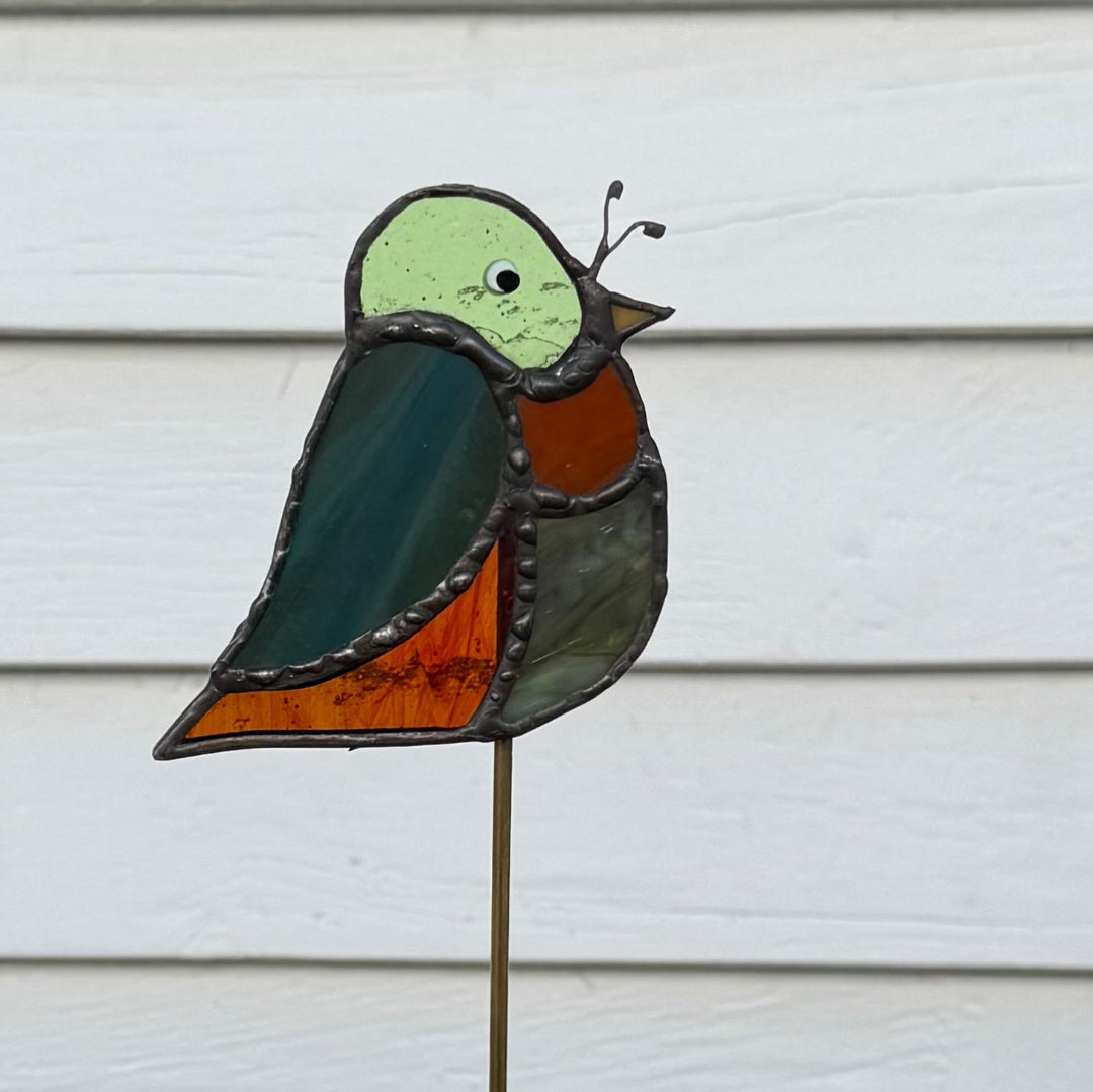 A stained glass bird garden stake featuring a light green head, dark teal wing, rust-orange chest, and gray body, mounted on a thin metal stake against a white siding background.
