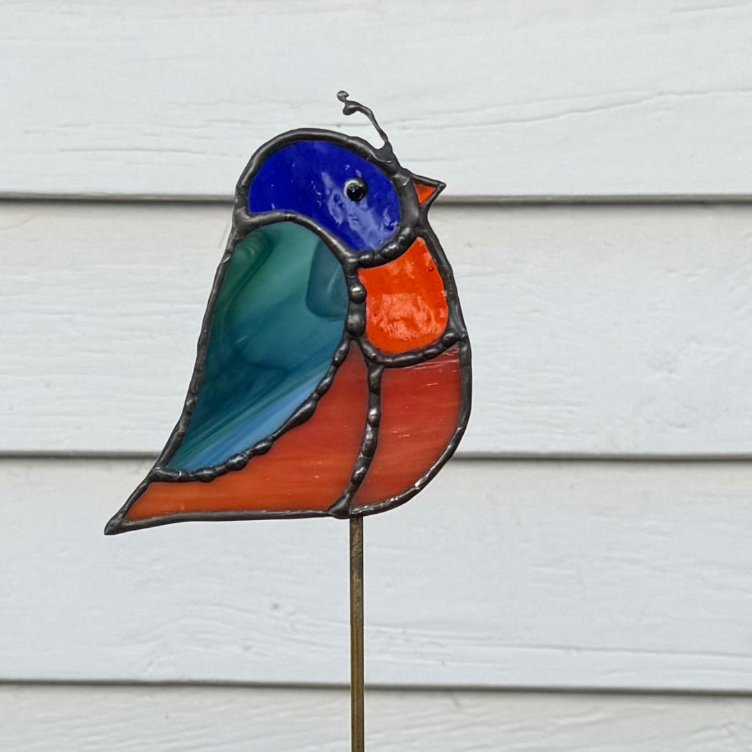 A stained glass bird garden stake featuring a bird design with a blue head, teal wing, orange chest, and reddish-orange body, mounted on a metal stake against a white siding background.
