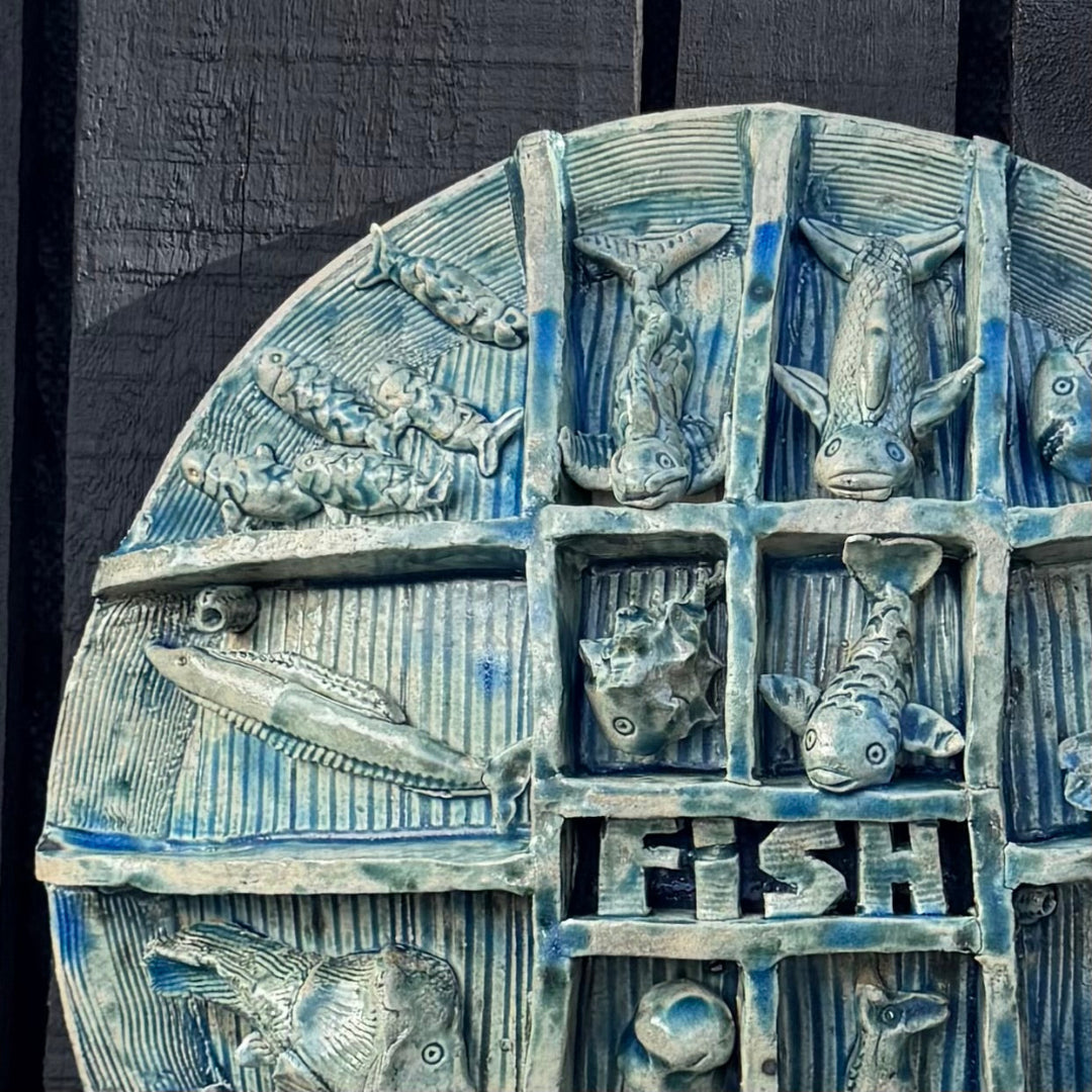 A close-up of a round decorative wall plaque featuring embossed fish motifs arranged in a grid with the word “FISH” at the center, finished in a blue-gray glazed ceramic or stone look against a dark background.