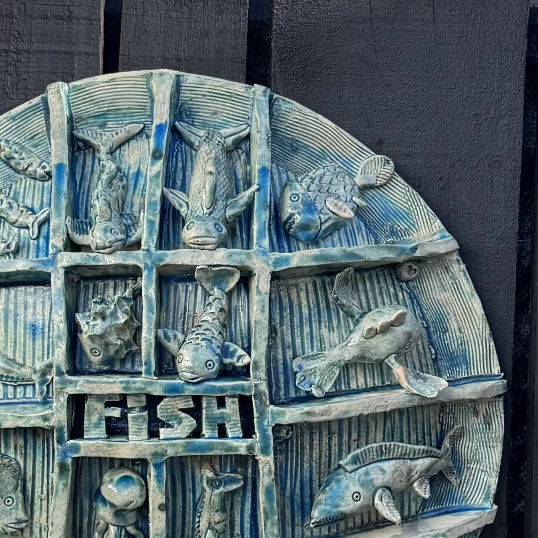 A round ceramic wall plaque featuring a grid of stylized fish in blue and white glaze, with the word 'FISH' cut through the center, mounted against a dark textured wall.
