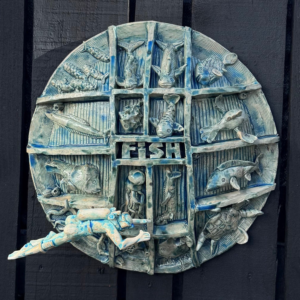 A round, multi-compartment ceramic wall sculpture featuring relief carvings of various sea creatures, including fish, a turtle, an octopus, a seahorse, and a scuba diver mounted in front on the lower left, set against a black background .