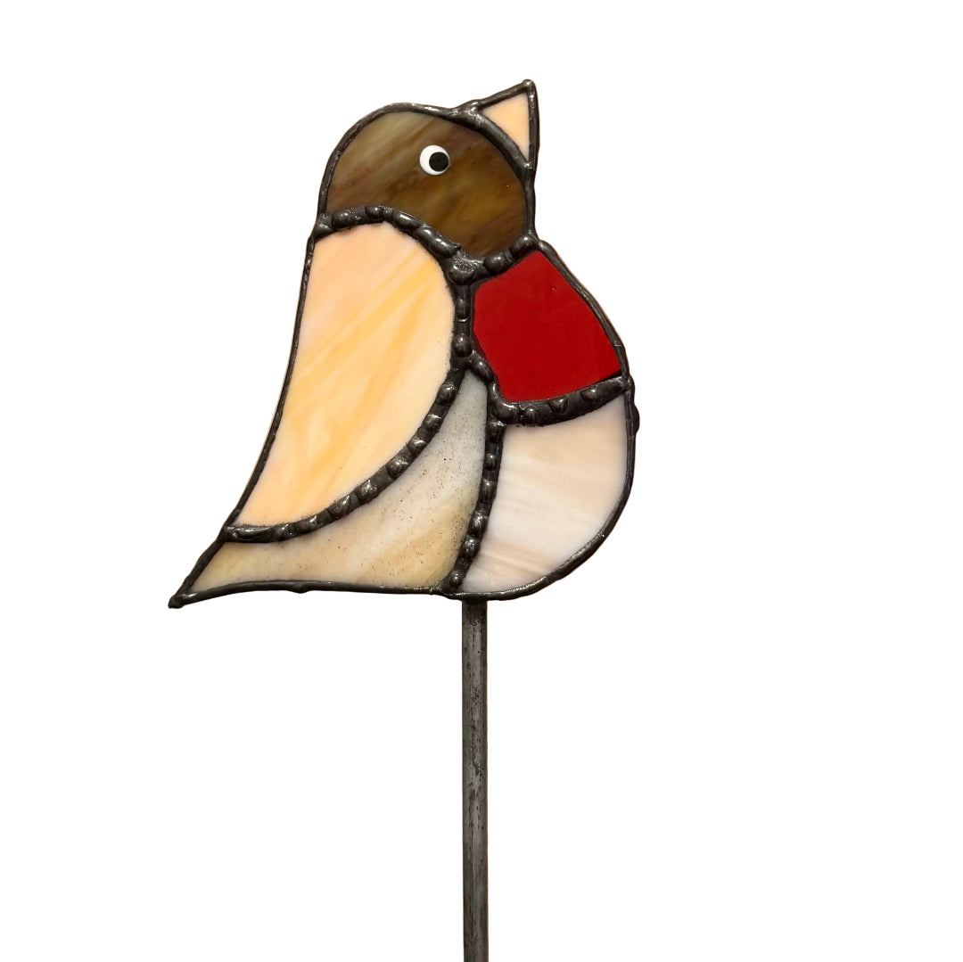 A stained glass bird ornament on a metal stake, featuring a brown head, cream and beige wings, a bright red chest, and dark metal leading, set against a plain tan background.