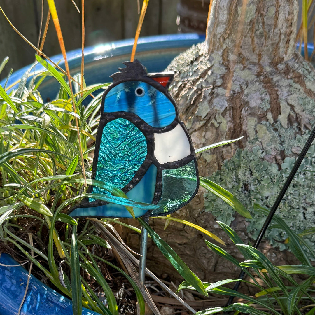 A stained glass bird garden stake with a blue, teal, and white design, displayed outdoors in green grass next to a tree trunk.