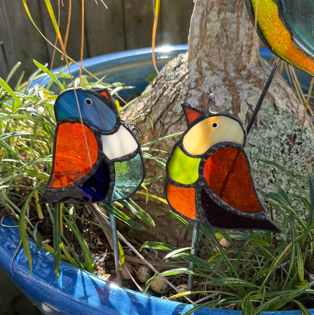 Two stained glass bird garden stakes with colorful abstract designs, placed in green grass next to a tree trunk in a blue planter.