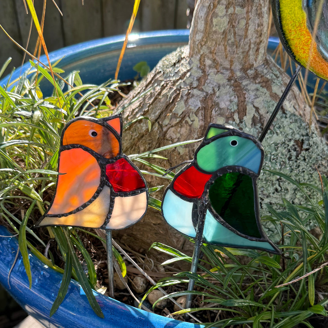 Two stained glass garden stakes shaped like birds, one orange and red and one green, red, and light blue, placed in a blue planter with grass and a tree trunk in the background.