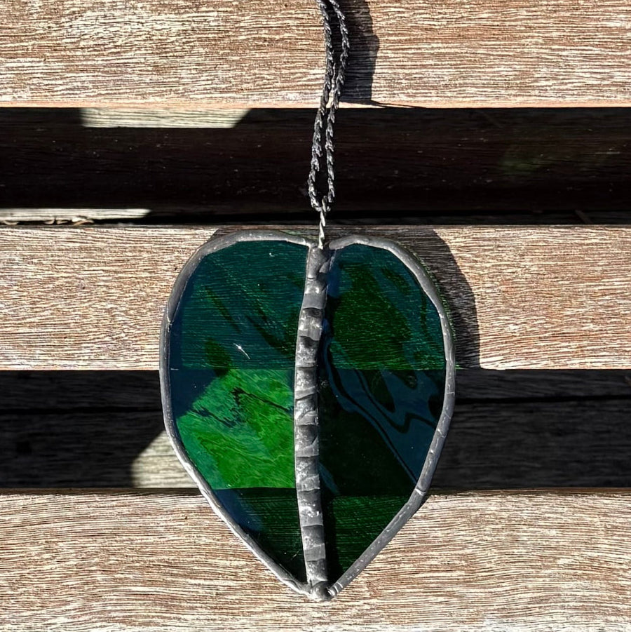 A Kawakawa Leaf stained glass ornament with clear green glass, set in a silver-colored metal frame and hanging on a rustic wooden slatted surface.