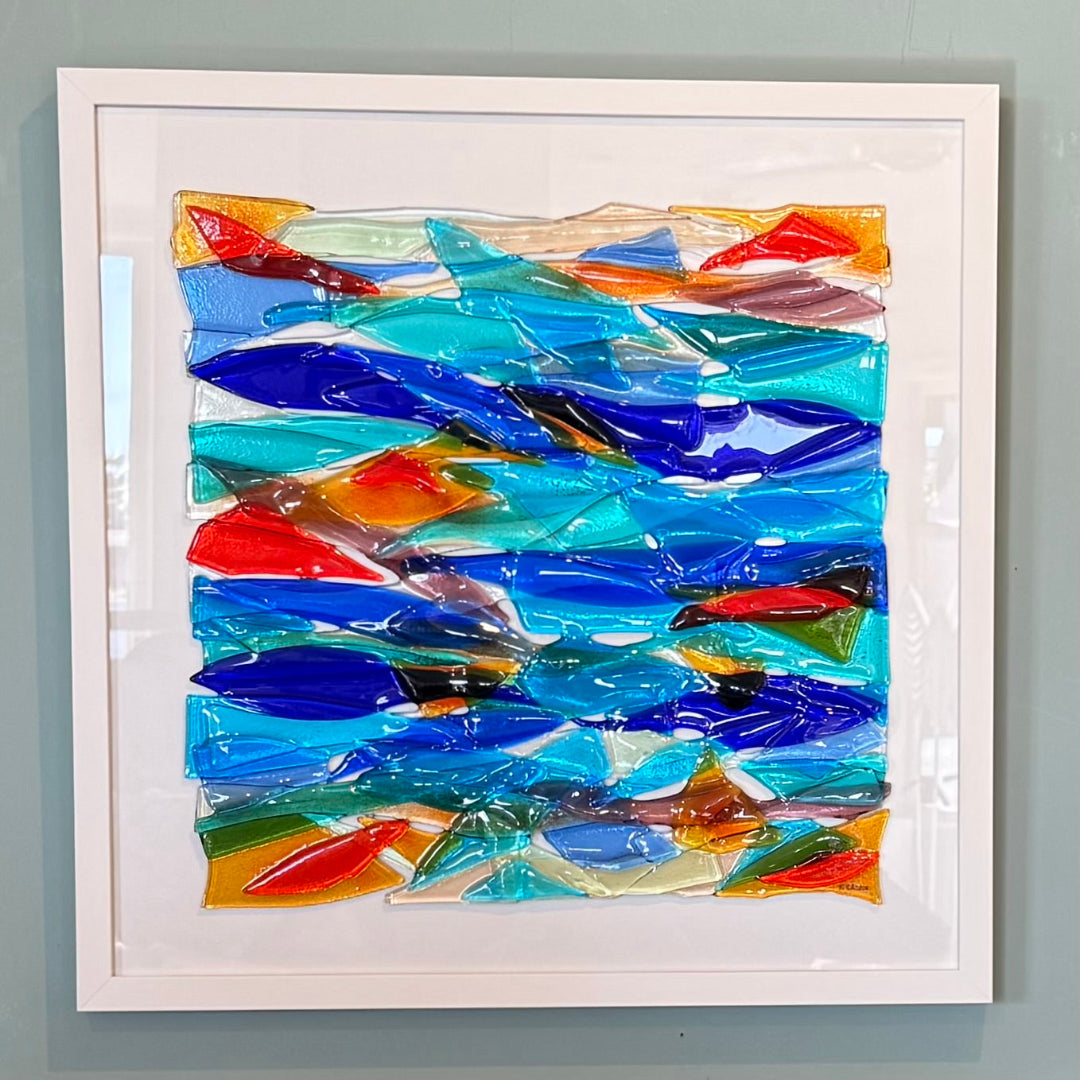A square abstract glass artwork with layered translucent blue, turquoise, red, orange, green, and brown shapes, presented in a white frame against a light gray wall.