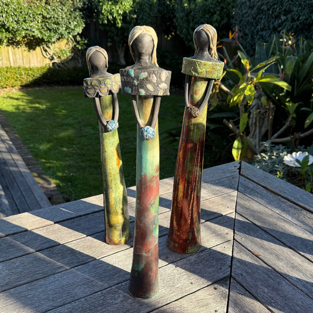 Three decorative standing figurines shaped like stylized women, displayed outdoors on a wooden deck with a garden background.