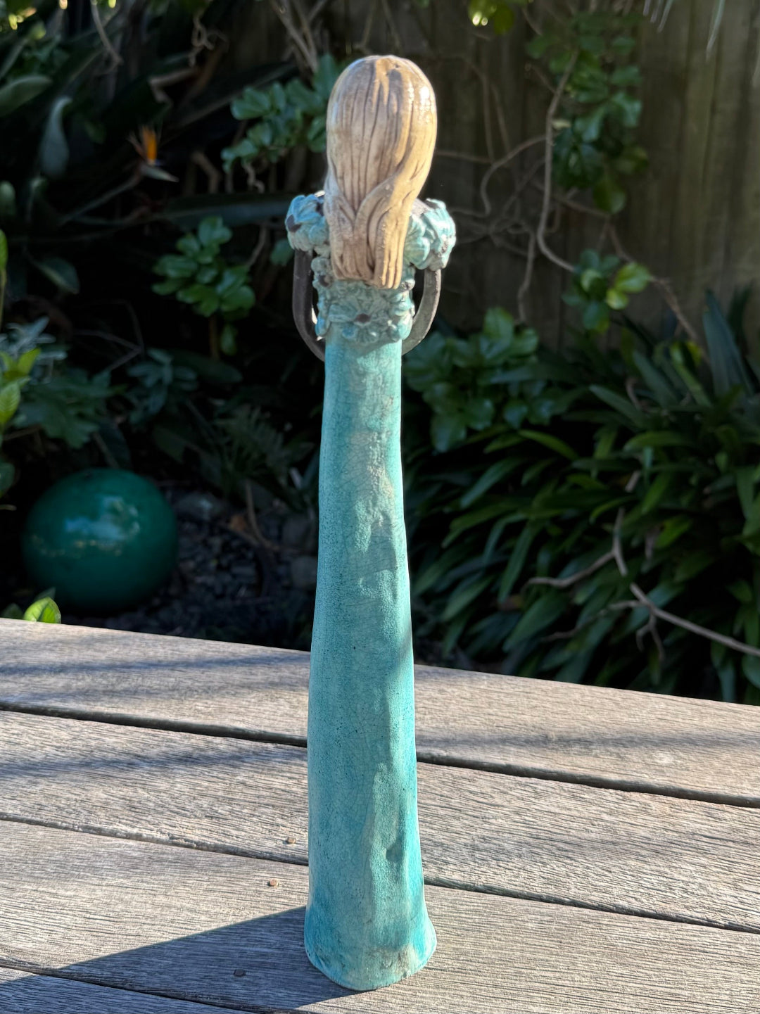 A decorative garden statue of a woman with long blonde hair and a turquoise patina body, standing on a weathered wooden deck with garden foliage in the background.