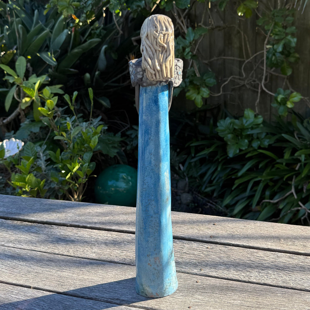 Back view of a decorative statue of a female with a blue body and a textured blond hair, standing on a wooden table in a garden setting.