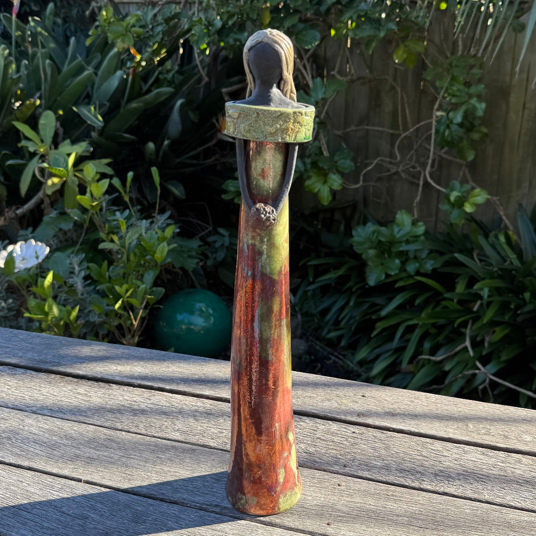 A tall, slender decorative ceramic figurine of a woman with long hair, wearing a green and rust-colored dress, standing on a weathered wooden deck with a lush garden background.