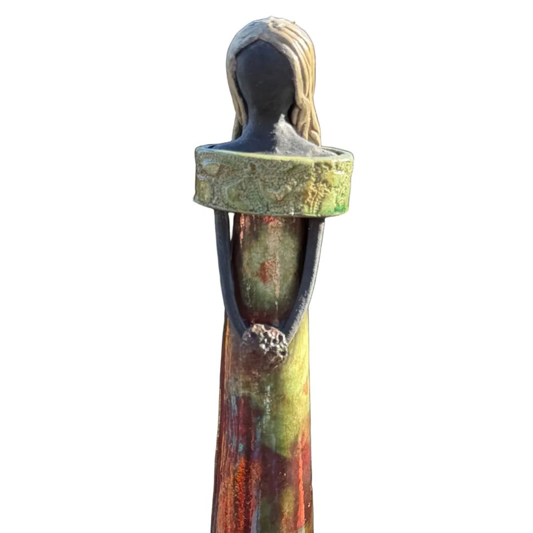 Close up view of a tall, slender decorative ceramic figurine of a woman with long hair, wearing a green and rust-colored dress with a white background.