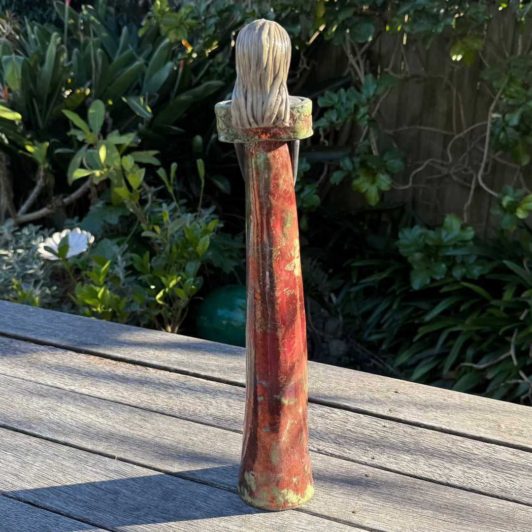 A back view of a tall, slender decorative ceramic figurine of a woman with long hair, wearing a green and rust-colored dress, standing on a weathered wooden deck with a lush garden background.