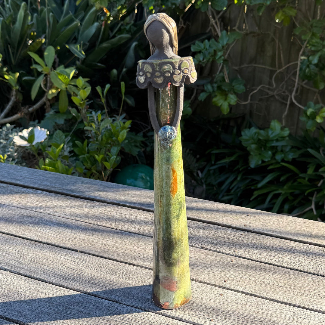 A tall, slender decorative statue with a stylized female figure, standing on a weathered wooden deck with a lush green garden background.