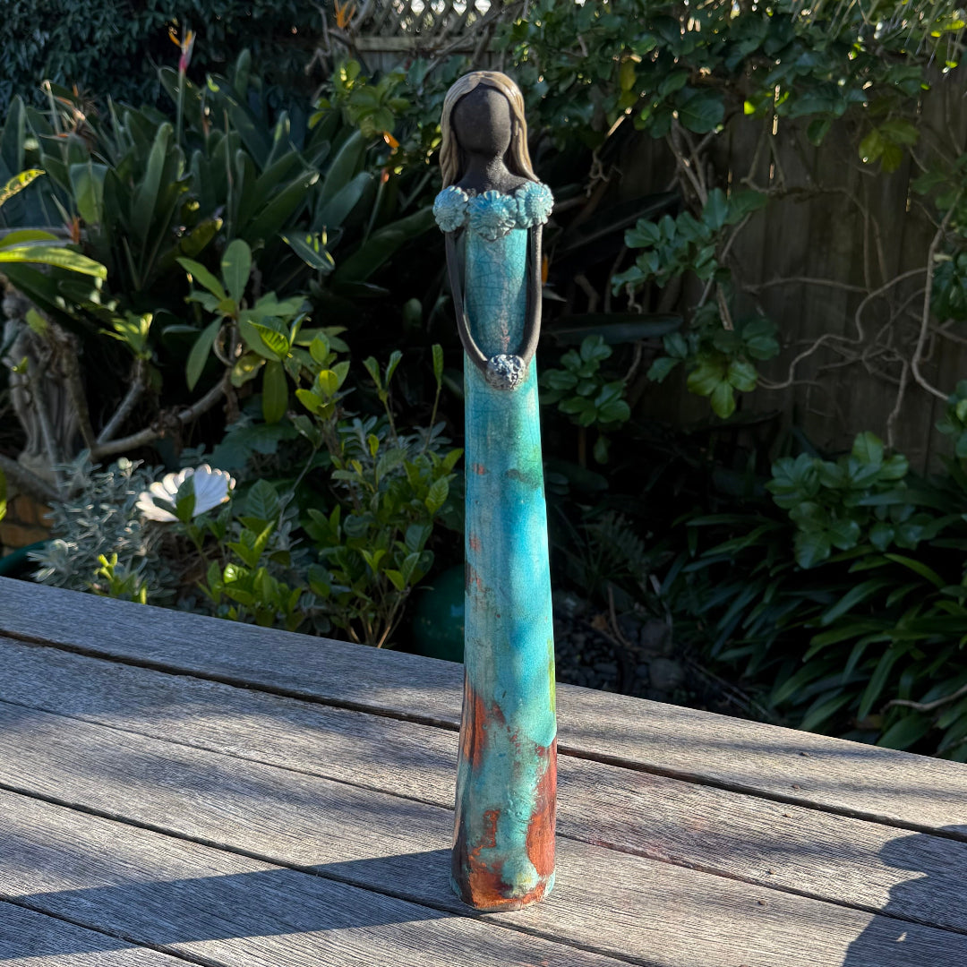 A tall, slender decorative figurine of a woman with long dark hair, wearing a turquoise and rust-colored dress, standing on a weathered wooden deck with a lush garden background.