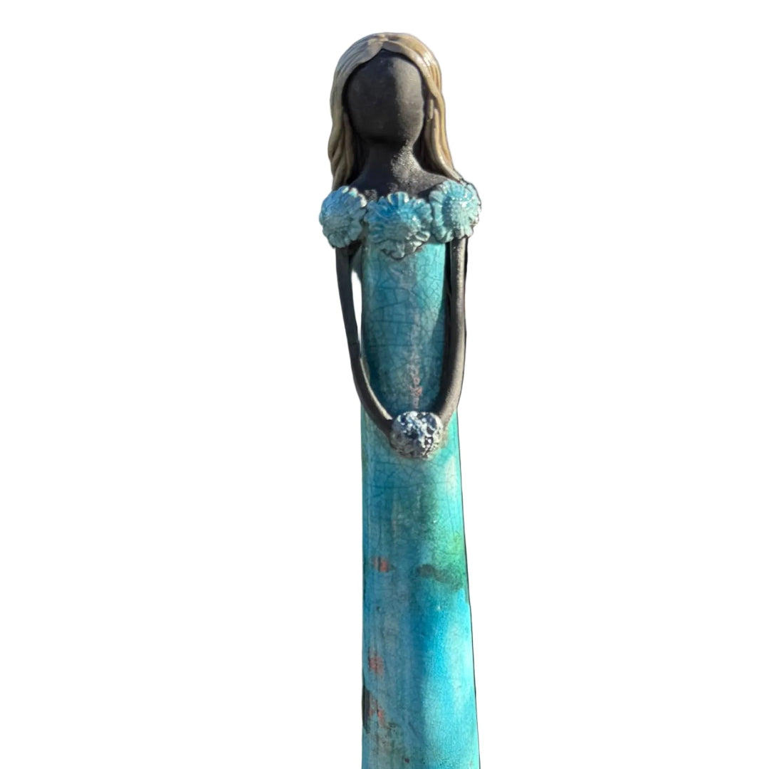 A close up view of a tall, slender, ceramic decorative figurine of a woman with long dark hair, wearing a turquoise and rust-colored dress, on a white background.