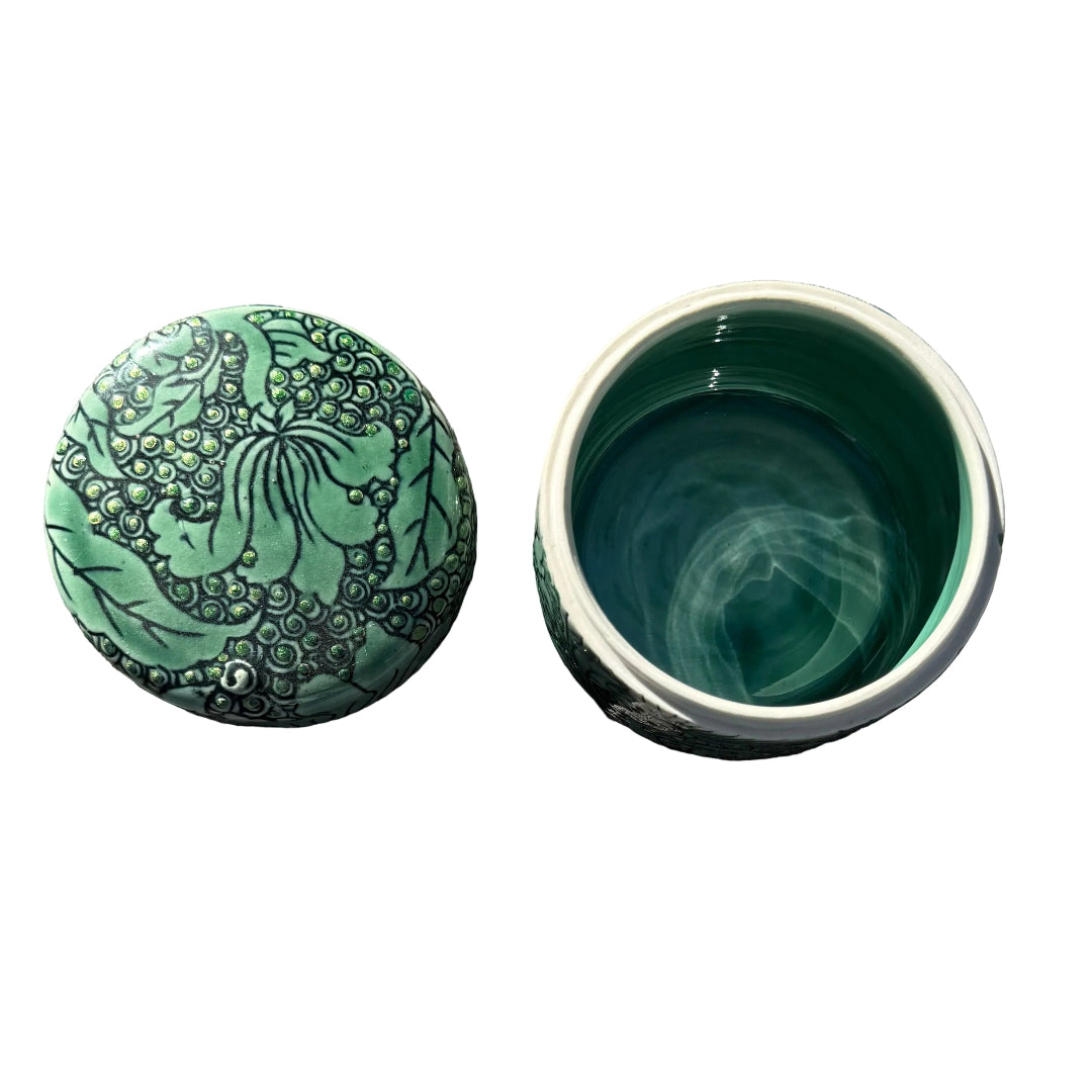 A green ceramic cylinder with a floral lotus pattern lid sits next to an open matching base in the sun on white surface.