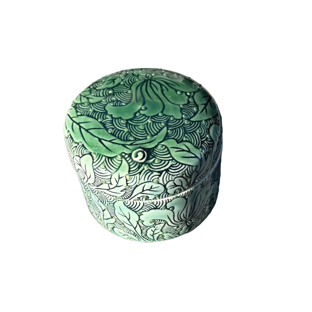 A small cylindrical ceramic box with a lid, decorated with a raised dots & a green and black lotus pattern, sitting on a white surface.