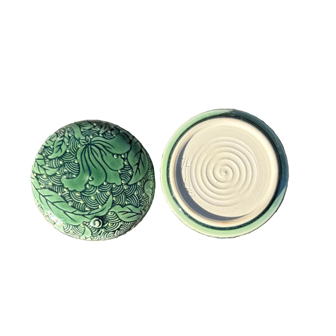 A small round ceramic container with its lid off, placed on a white surface. The lid is green with carved floral lotus patterns, while the bottom of the container shows a cream-colored interior with concentric spiral ridges and a green rim signed with the initials S/L.