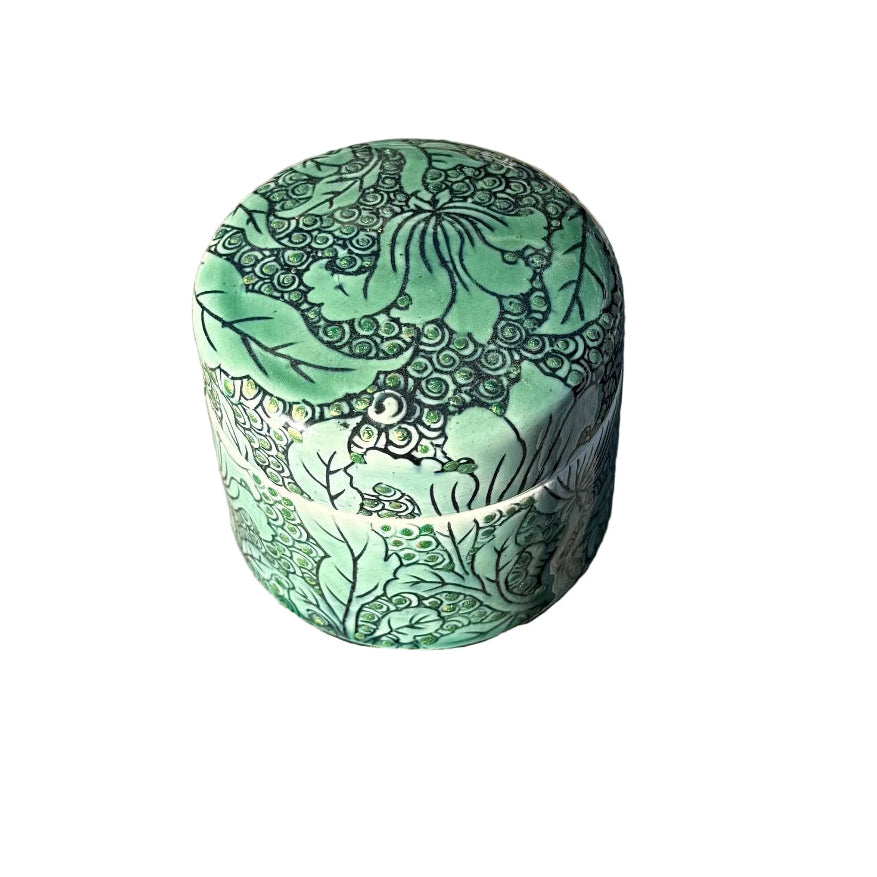 A small cylindrical ceramic box with a lid, decorated with a green and black lotus pattern, sitting on a white surface.