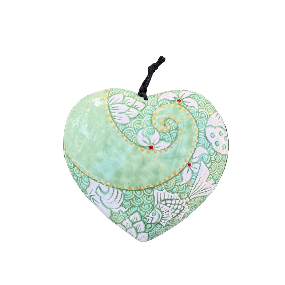 A heart-shaped decorative wall hanging with a black hanging string, featuring a light green and white floral pattern, displayed on a white wall.