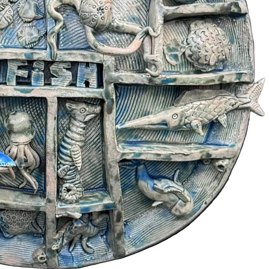 Close up view of a round, multi-compartment ceramic wall sculpture featuring relief carvings of various sea creatures, including fish, a turtle, an octopus, a seahorse, and a shark, with a blue and white shark mounted in front on the lower left, set against a white background .
