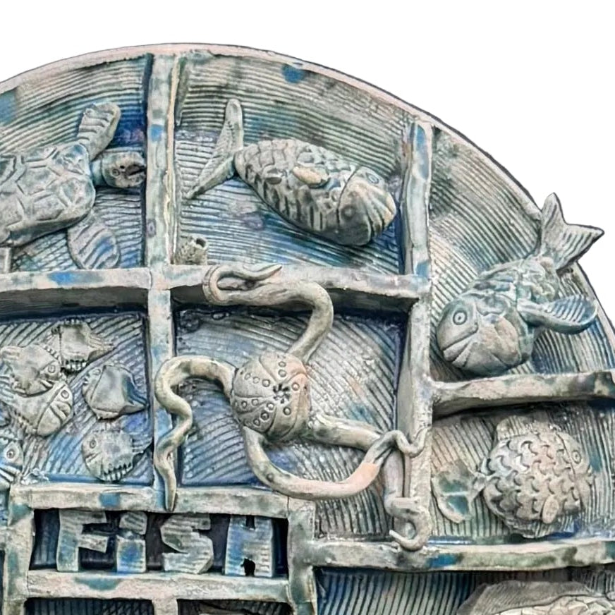Close up view of a round, multi-compartment ceramic wall sculpture featuring relief carvings of various sea creatures, including fish, a turtle, an octopus, a seahorse, and a shark, with a blue and white shark mounted in front on the lower left, set against a white background .
