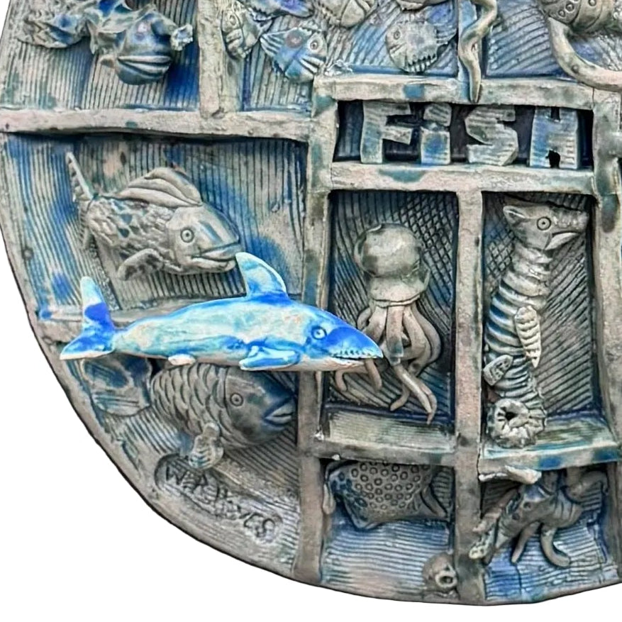 Close up view of a round, multi-compartment ceramic wall sculpture featuring relief carvings of various sea creatures, including fish, a turtle, an octopus, a seahorse, and a shark, with a blue and white shark mounted in front on the lower left, set against a white background .
