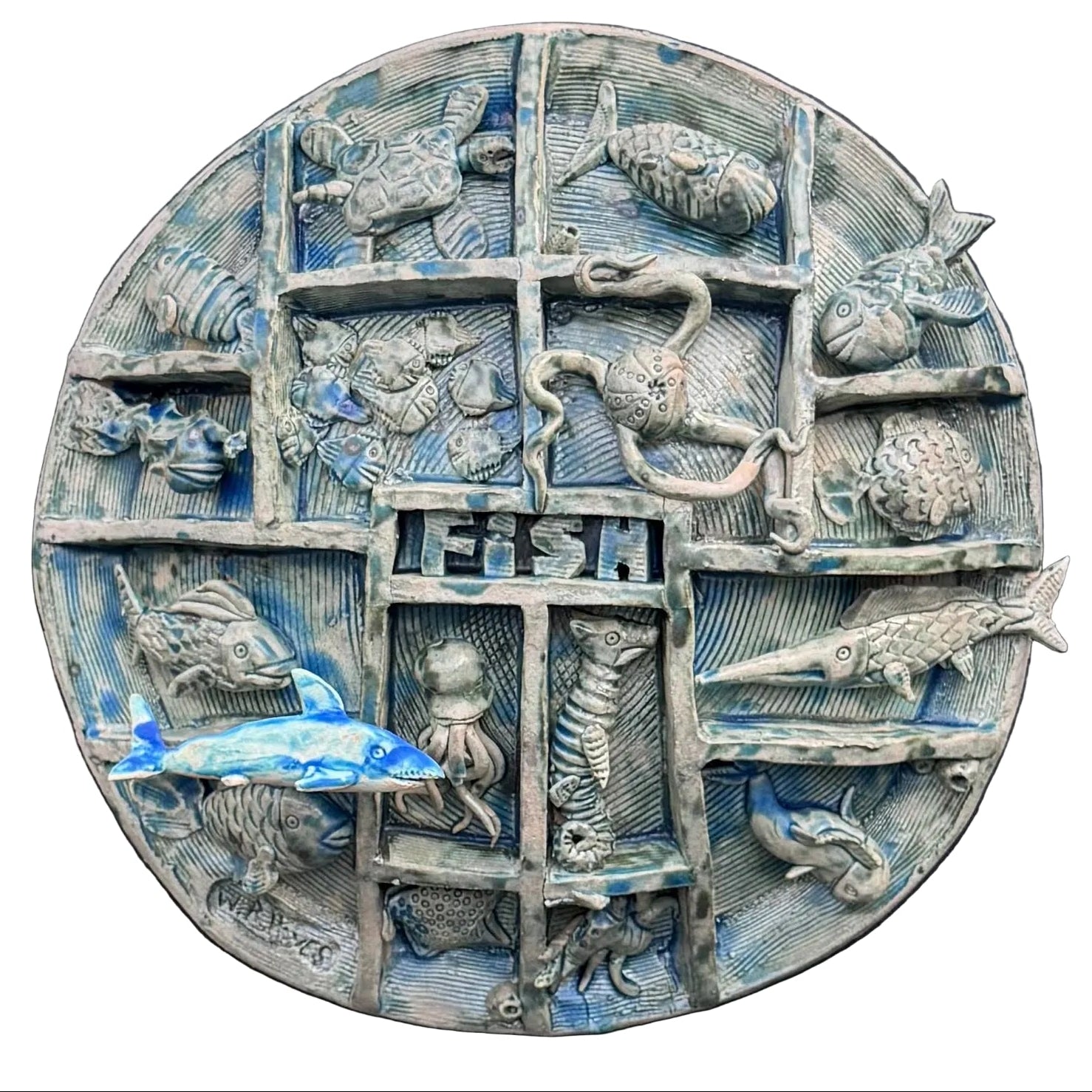 A round, multi-compartment ceramic wall sculpture featuring relief carvings of various sea creatures, including fish, a turtle, an octopus, a seahorse, and a shark, with a blue and white shark mounted in front on the lower left, set against a white background .