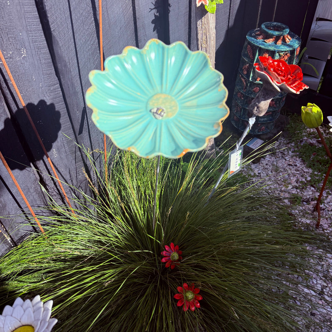A cluster of decorative garden metal flower sculptures in various colors and styles, including a mint green daisy with a bee, white daisies, and red blossoms, arranged in front of a dark gray wooden fence.