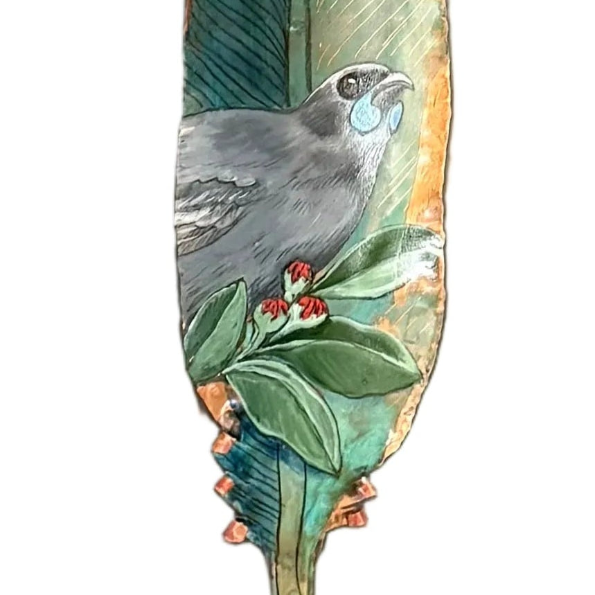 A close up view of a hand-painted Kokako bird on a copper feather wall sculpture, painted with green, blue, and red details, on a white background.