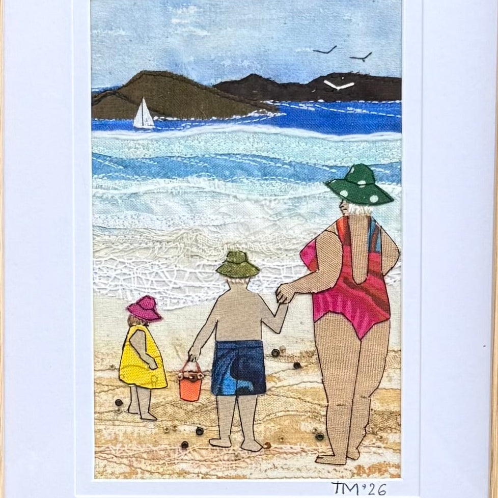 A close up view of a small each textile artwork framed with a white mat. The artwork depicts a beach scene with a woman & 2 children playing.