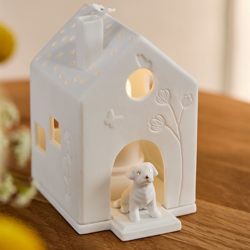 A white ceramic dog house candle holder with a small white dog figurine inside, placed on a wooden surface with warm light glowing from within.