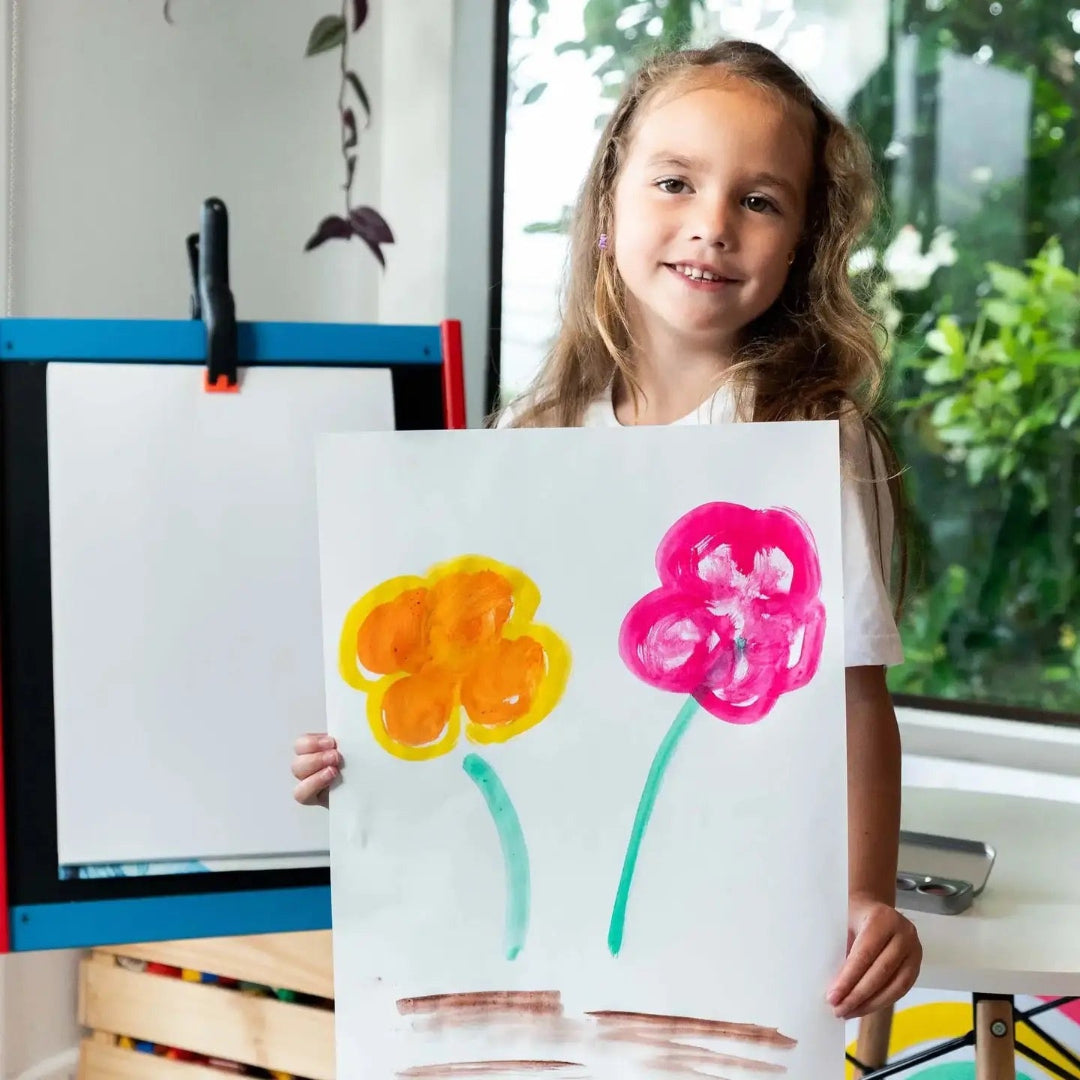 Child holding a drawing of two flowers, one yellow and one pink, in an indoor setting.