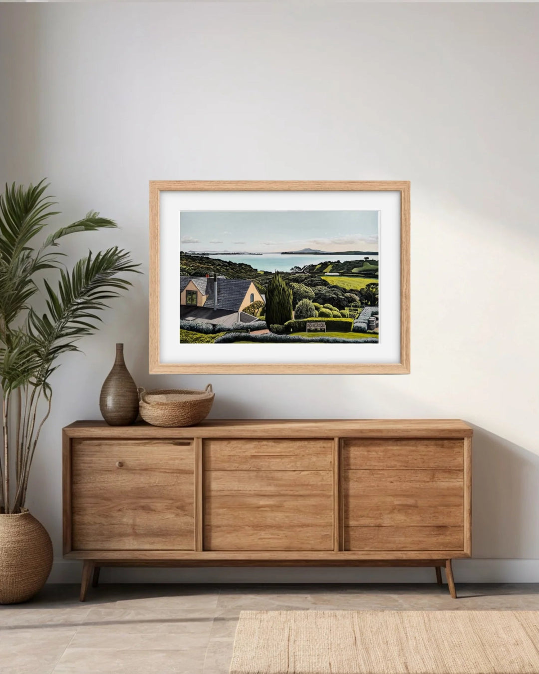 A framed landscape art print depicting a coastal village with green fields and a blue sea, styled on a light gray wall above a mid-century modern wooden sideboard with a potted plant and decorative vases.