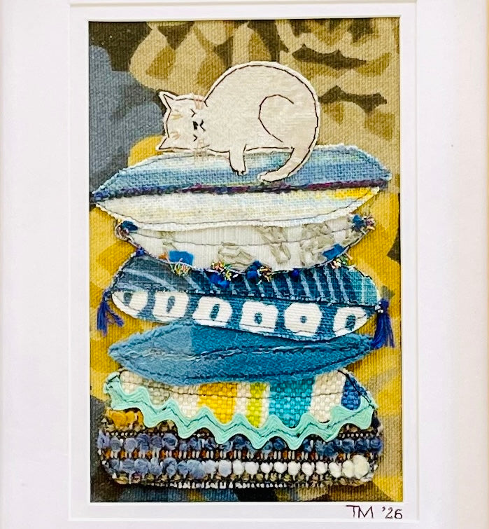 Framed textile art featuring a white cat curled up on top of a stack of blue and white pillows, with a yellow patterned background.